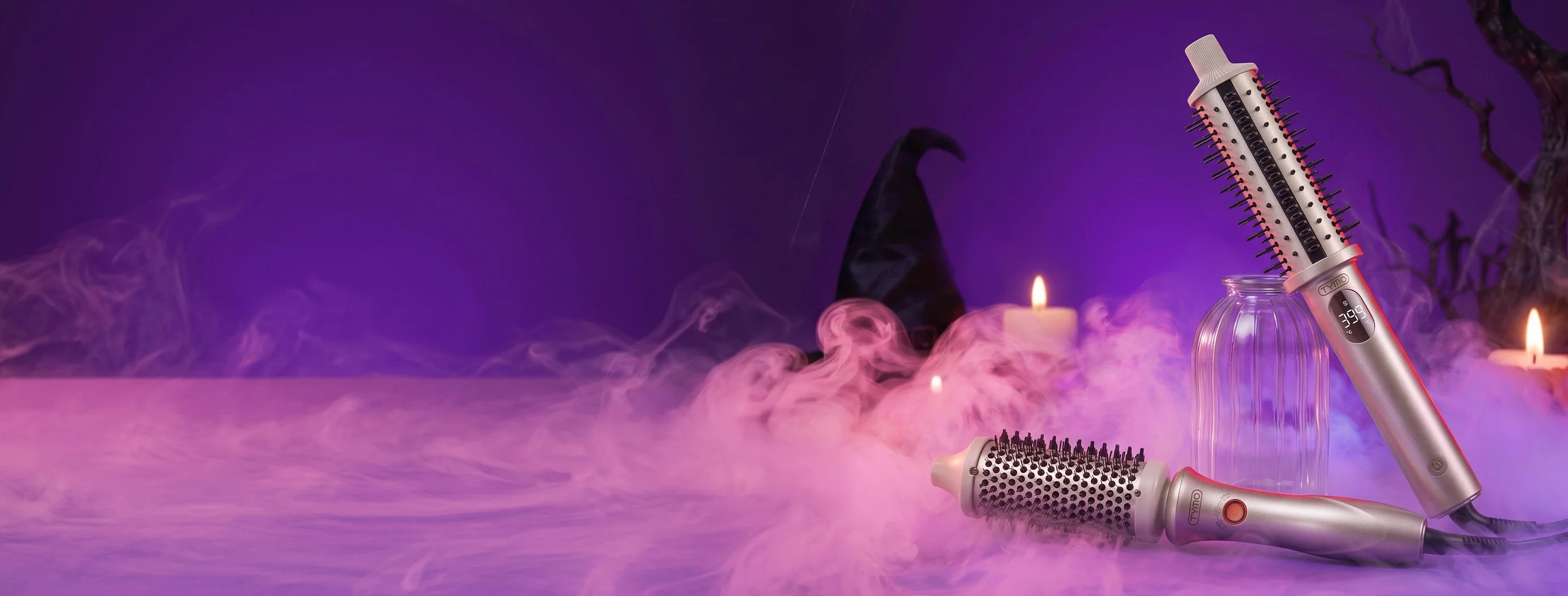 Two silver and white hot air styler tools with bristles, styled on a purple surface with pink fog, a witch hat, candles, and a glass jar in a spooky Halloween setting.
